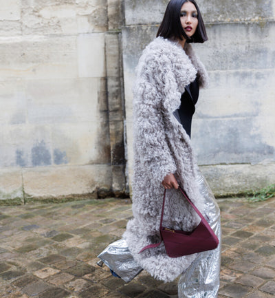 A woman wearing a shearling trench coat in grey, silver pants and heels, holding an aubergine italian leather Lulu handbag from Studio DRÈA for a L'Officiel Monaco editorial photo shooting