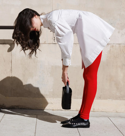 A model wearing red tights, a white chemise, and black ballerina shoes holding the Studio DRÈA Violette black leather handbag in a L'Officiel  editorial photo shooting